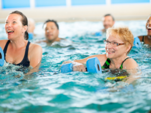 Smiling older adults using foam dumbbells during an aqua aerobics session in the WynActive pool.