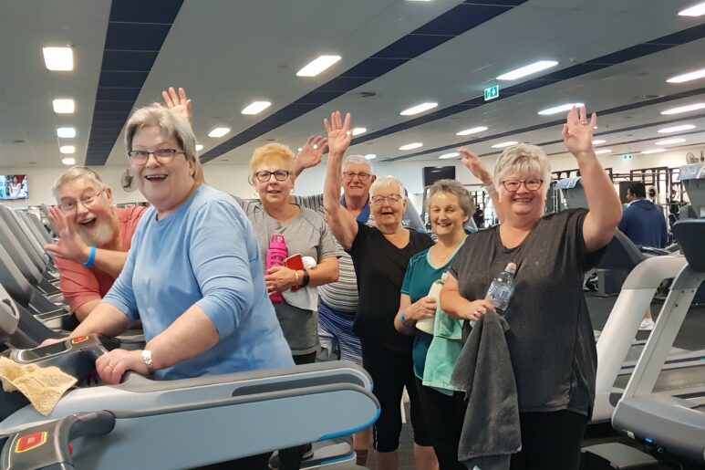 Group of older adults smiling and waving while exercising on treadmills at WynActive gym