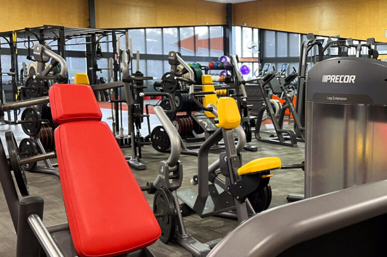 Gym equipment area with a red-seated weight machine and multiple strength machines arranged near large windows.