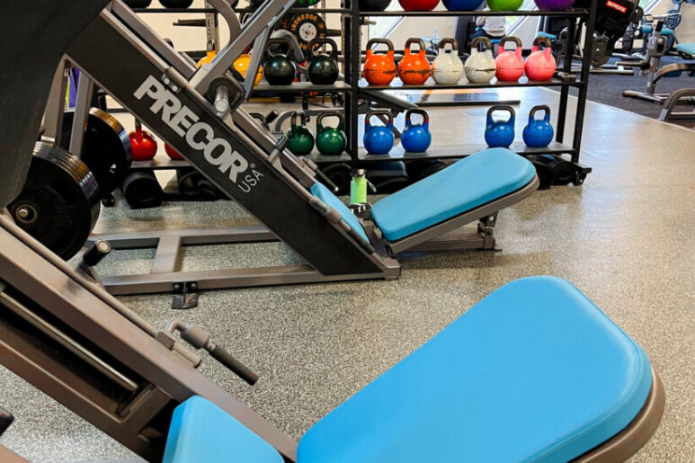 Leg press machine with bright blue seating in front of racks filled with colourful kettlebells, medicine balls, and other gym equipment.