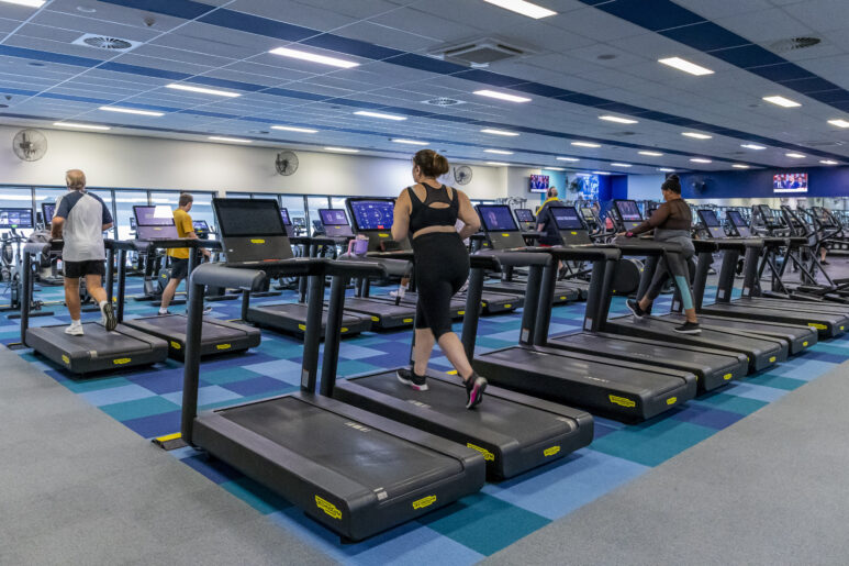 People exercising on treadmills inside a large gym with rows of cardio machines, bright lighting, and blue patterned flooring.