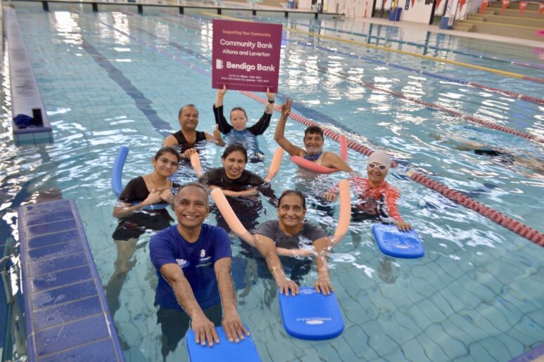 Group of adults in a swimming pool at AquaPulse Hoppers Crossing holding kickboards and pool noodles, smiling for a class photo.
