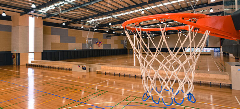 Close-up view of a basketball hoop inside Eagle Stadium, Wyndham, overlooking polished timber indoor courts lined for basketball and other sports.