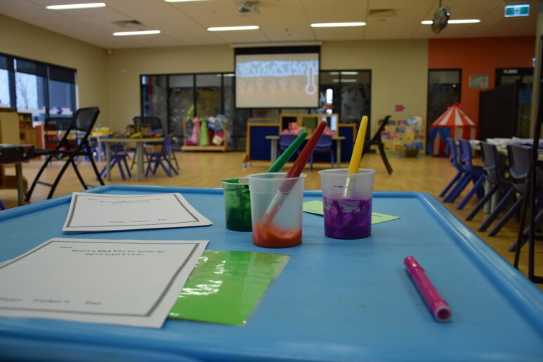 Table set up with paint pots and brushes in a childcare craft area, with toys and play stations in the background.
