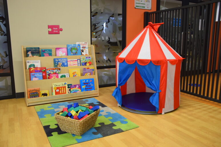 Reading corner with children’s books, a basket of foam blocks, and a red-and-white play tent.