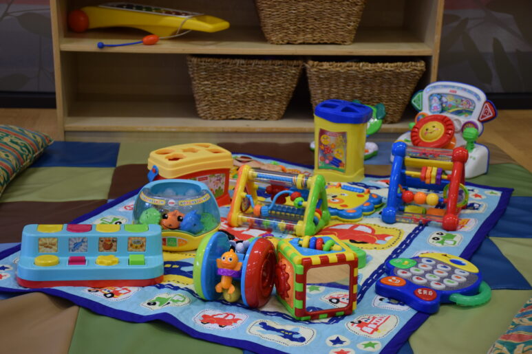 Assortment of colourful baby toys and activity centres laid out on a padded play mat in a childcare room.
