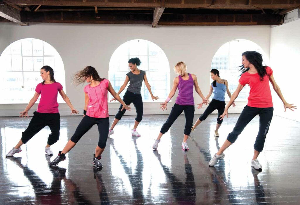 Women in a dance class performing choreographed moves together on a polished wooden floor in a loft-style studio.