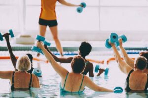 Group aqua fitness class with participants lifting foam dumbbells, led by an instructor poolside.