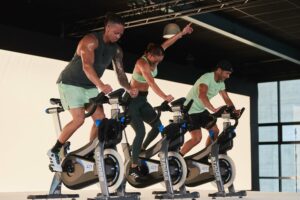 Three instructors on stage leading a high-energy spin class, riding stationary bikes in unison.