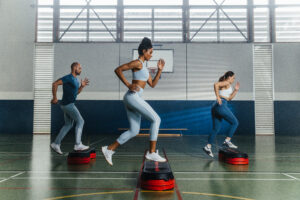 Group fitness class stepping on platforms in a sports hall, performing cardio step exercises.