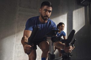 Man in a blue shirt riding a stationary bike during a spin session, with another participant blurred in the background.