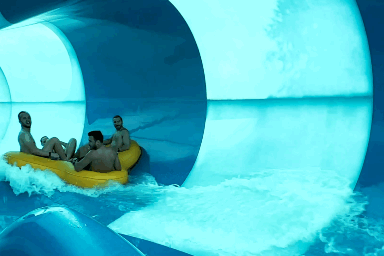 Group of people riding a yellow inflatable raft down the blue waterslide at AquaPulse Hoppers Crossing, splashing into the pool at the slide’s exit.