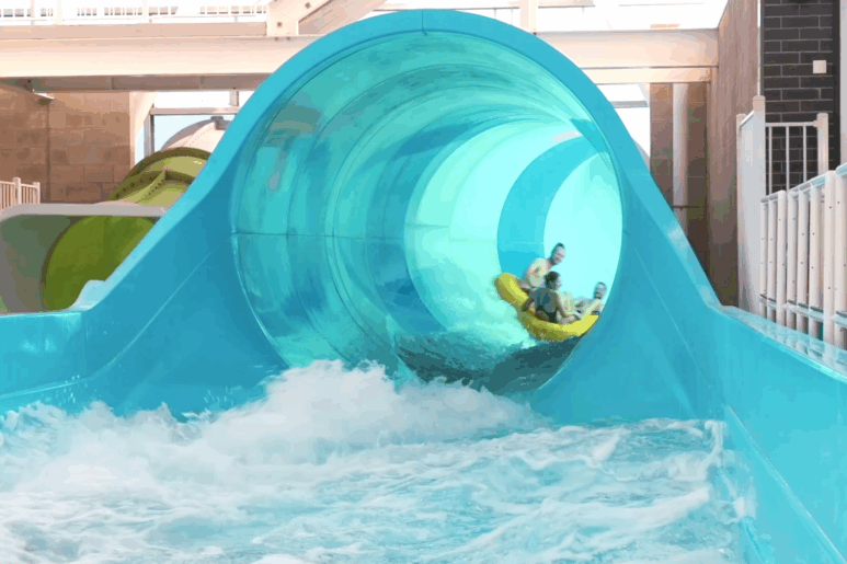 Group of people riding a yellow inflatable raft down the blue waterslide at AquaPulse Hoppers Crossing, splashing into the pool at the slide’s exit.