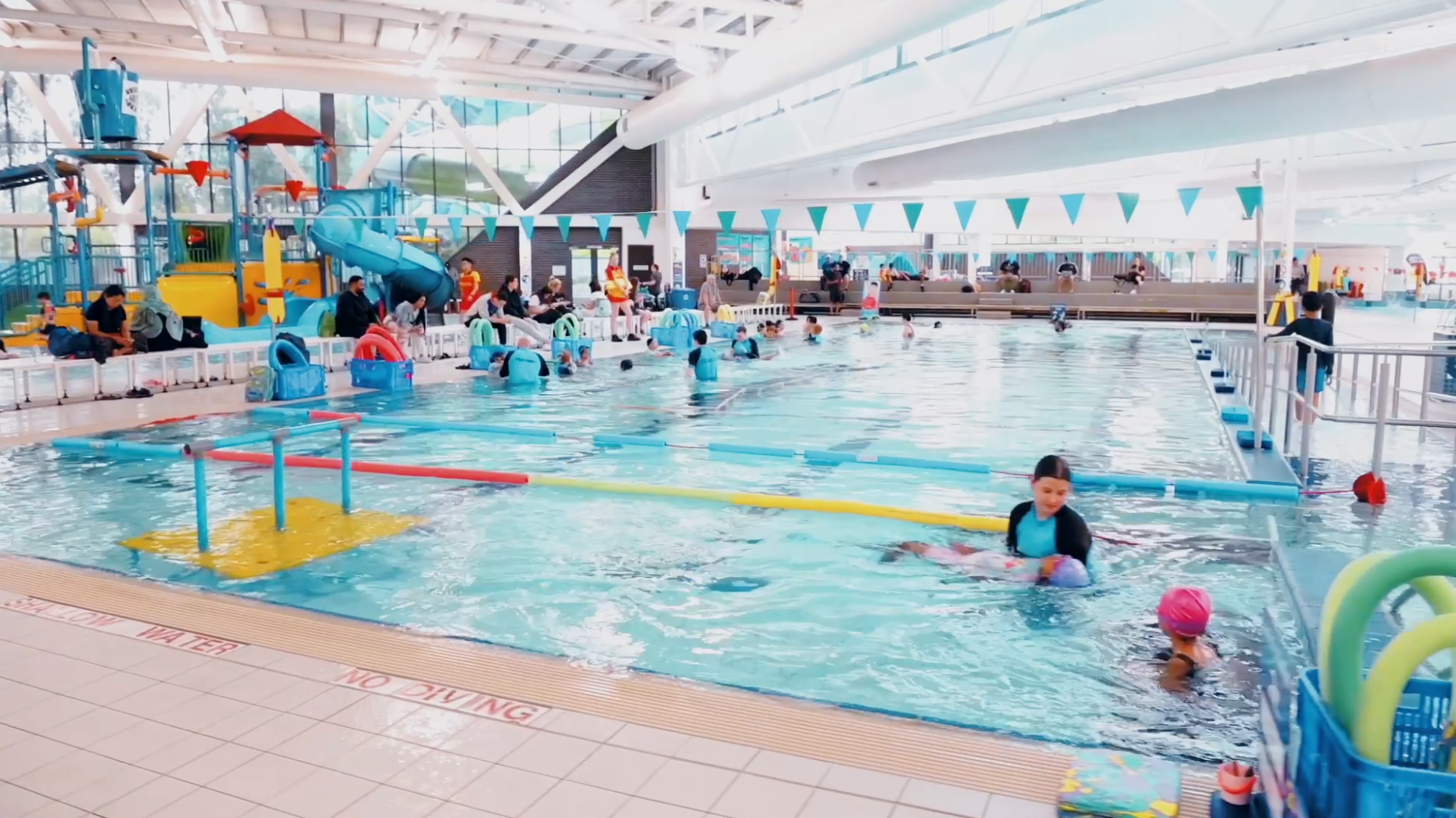 Indoor swimming pool at AquaPulse Hoppers Crossing with children in swimming lessons, instructors in blue uniforms, and colourful play structures in the background.
