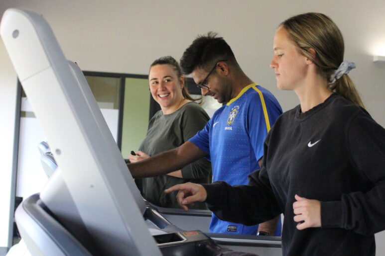 Three people walking on treadmills, one smiling at the camera during a gym session.