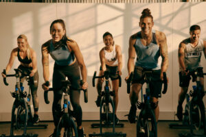 Group of five people in a spin class, smiling and motivated while cycling indoors.