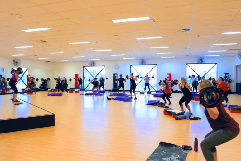 Large group fitness class with participants lifting barbells during a BodyPump session in a bright studio with mirrors, wooden floors, and exercise steps.