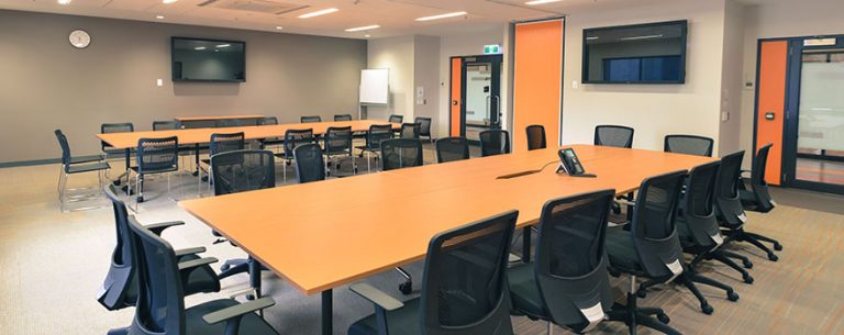 Modern meeting room at Eagle Stadium with long conference tables, black chairs, wall-mounted screens, and bright orange accents.