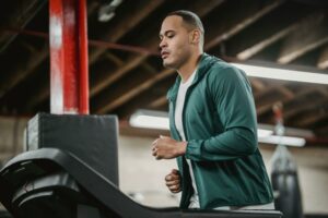 Man in a green jacket running on a treadmill in a gym with red beams.